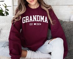 a woman sitting on top of a couch wearing a maroon grandma sweatshirt with white lettering