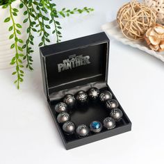 a black box with silver balls in it on a white table next to some plants