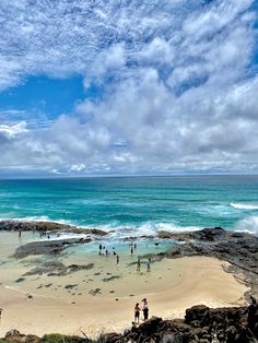 several people are standing on the beach near the water and rocks, while others stand in the sand