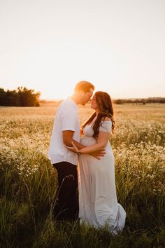 a pregnant couple standing in the middle of a field at sunset with their arms around each other