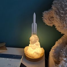 a white teddy bear sitting next to a lit up rocket ship on top of a table