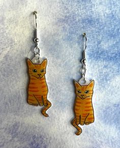 two orange cats sitting on top of each other dangling from silver earwires in front of a blue background