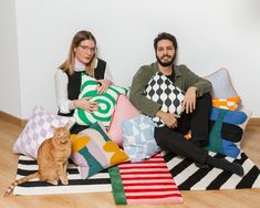 two people sitting on the floor with pillows and a cat