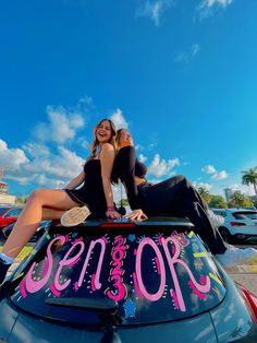 two women sitting on top of a car with the word senior spelled in pink letters