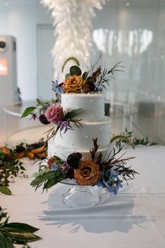 a three tiered wedding cake with flowers and greenery on the table in front of a chandelier