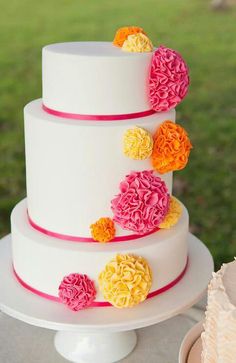 a three tiered white cake with pink and yellow flowers on the top, sitting on a table outside