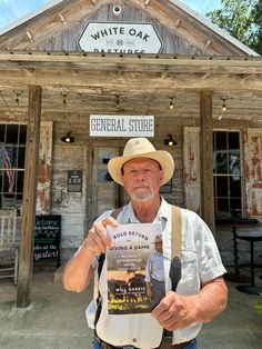 an older man wearing a hat and suspenders holding up a book in front of a building