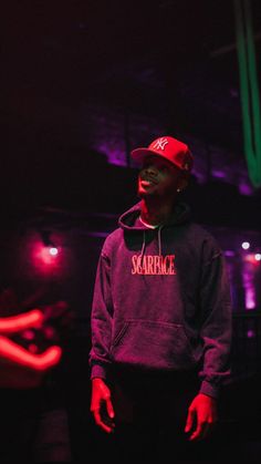 a young man wearing a red hat and black hoodie in a dark room with neon lights