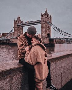 a man and woman sitting next to each other on a wall near the water with a bridge in the background