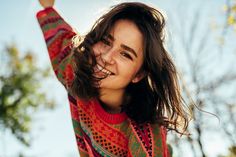 a woman with long hair wearing a colorful sweater smiles and holds her arms in the air