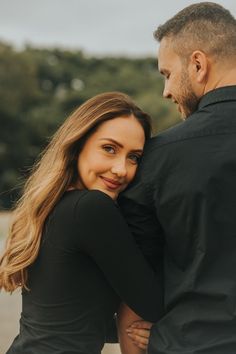 a man and woman embracing each other in front of the camera, with trees in the background