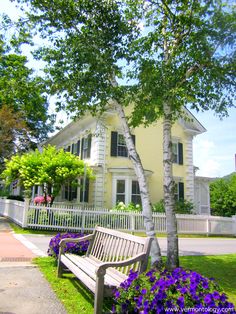 a wooden bench sitting in front of a yellow house with purple flowers growing around it