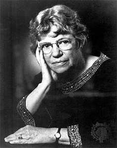 an old black and white photo of a woman with glasses sitting at a table looking off to the side