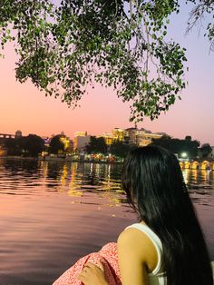 a woman sitting on the edge of a body of water looking at the city lights