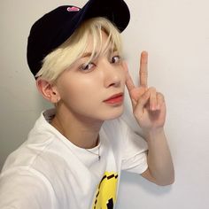 a person with blonde hair wearing a baseball cap and making the peace sign while standing against a wall