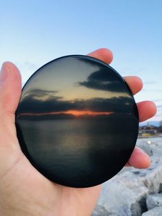 a hand holding a round object in front of the ocean at sunset, with clouds reflected on it