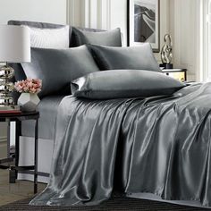 a bed with grey sheets and pillows in a room next to a table, mirror and lamp