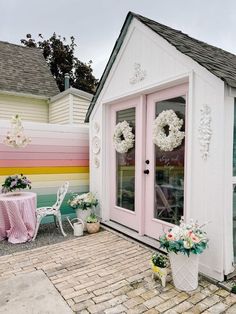 a small white house with pink doors and flowers on the front door, next to a brick patio