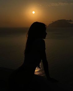 a woman sitting on the edge of a cliff watching the sun go down over the ocean