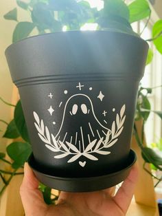 a hand holding up a black pot with white designs on the bottom and sides, in front of a plant