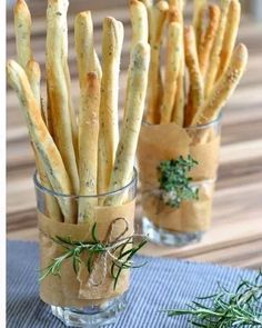 some bread sticks are in a small glass cup on a table with rosemary sprigs