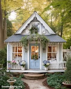 a small white house with flowers on the front door and porch, surrounded by greenery