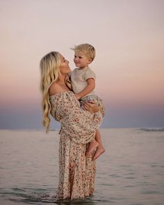 a woman holding a small boy in her arms while standing on the beach at sunset