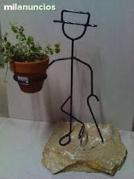 a wire man holding a potted plant on top of a rock next to a wall