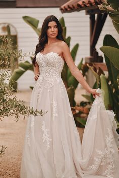 a woman in a white wedding dress standing next to some plants and holding a bag