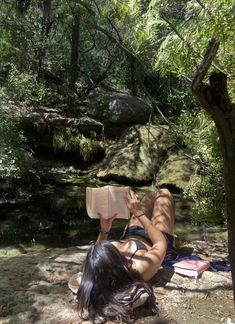 a woman reading a book while laying on the ground next to a river and trees