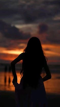 a woman standing on top of a beach next to the ocean at sunset with her arms behind her back