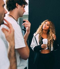 a woman standing in front of a mirror holding a cup and talking to another man