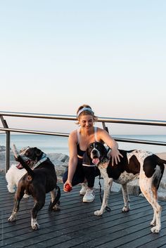 a woman kneeling down next to three dogs on a boardwalk near the ocean and water