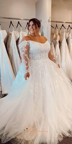 a woman in a white wedding dress standing next to dresses on hangers and looking at the camera