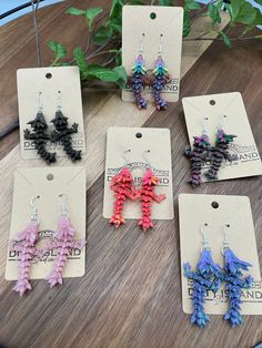 six pairs of earrings are displayed on a wooden table next to a plant with green leaves