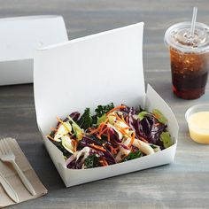 a salad in a white box next to a cup of juice and spoons on a table