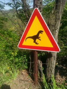 a yellow and red triangular sign with a dinosaur on it's side next to a tree