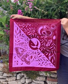 a woman holding up a purple and red quilt in front of some flowers on a stone wall