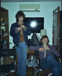 two people standing in a living room with one person holding a pipe and the other sitting on a chair