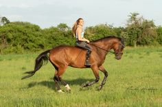 a woman riding on the back of a brown horse through a lush green field with trees in the background