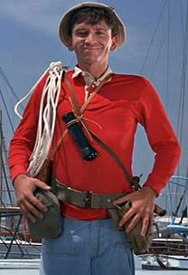 a man wearing a red shirt and hat standing in front of sailboats with his hands on his hips