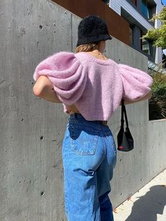 a woman with her back to the camera, wearing jeans and a pink sweater is leaning against a concrete wall