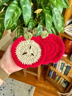 a hand holding two crocheted apples in front of a potted plant