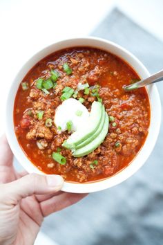 a person holding a bowl of chili with avocado and sour cream on top