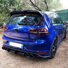 the rear end of a blue car parked in front of a fenced area with trees