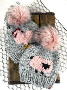 two knitted hats with pink pom - poms sitting on top of a piece of wood