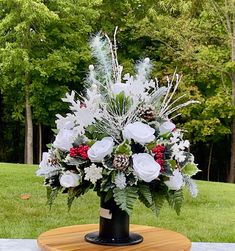 a black vase filled with white and red flowers on top of a wooden table in front of trees