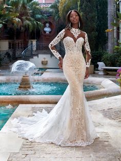 a woman standing in front of a fountain wearing a wedding dress with long sleeves and an open back