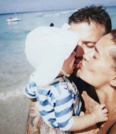 a man holding a baby on the beach with boats in the water behind him and his mother kissing him