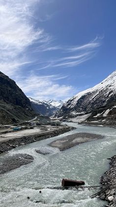 a river flowing through a valley surrounded by snow covered mountains under a blue sky with wispy clouds